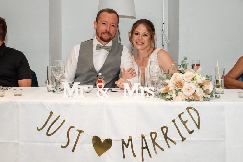 Hochzeitskrone Ein Paar im Hochzeitskleid sitzt an einem Tisch mit einem „Mr & Mrs“-Schild und einem „Just Married“-Banner und fängt bezaubernde Momente der Hochzeitsfotografie in Niederösterreich ein.