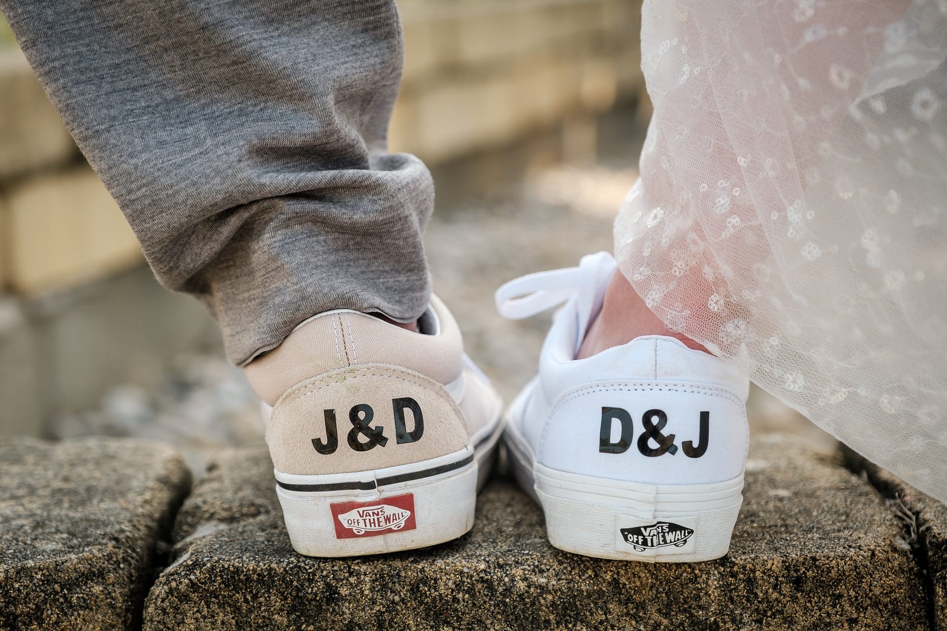 Hochzeitskrone Eine Nahaufnahme der Schuhe eines Paares zeigt die individuellen Initialen „J&D“ und „D&J“ auf den Absätzen, wobei eine der beiden graue Hosen und die andere ein weißes Kleid trägt und auf einer Steinoberfläche steht. Dieser bezaubernde Moment fängt die Essenz natürlicher Hochzeitsfotos ein.