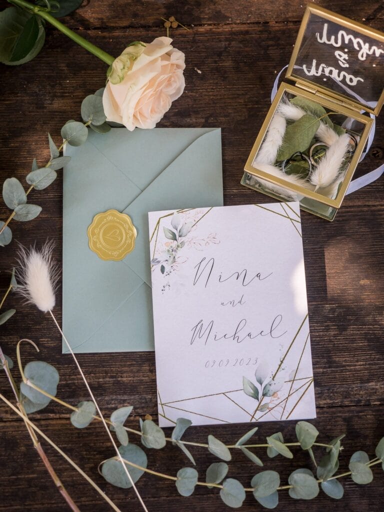 Hochzeitskrone Eine Hochzeitseinladungskarte liegt auf einem Holztisch neben einer zarten Rose, einem grünen Umschlag und dekorativem Eukalyptus. Eine kleine Glasbox umschließt die Ringe und hält bezaubernde Momente fest, perfekt für Hochzeitsfotografie in Niederösterreich.