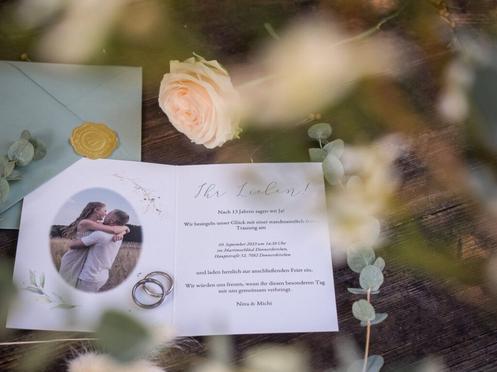 Hochzeitskrone Eine Hochzeitseinladung mit einem Foto des Paares, zwei Ringen und einer weißen Rose ziert eine Holzoberfläche neben einem grünen Umschlag. Der Text ist auf Deutsch und fängt bezaubernde Momente mit natürlicher Eleganz ein, die den Stil der Hochzeitsfotografie in Niederösterreich widerspiegelt.